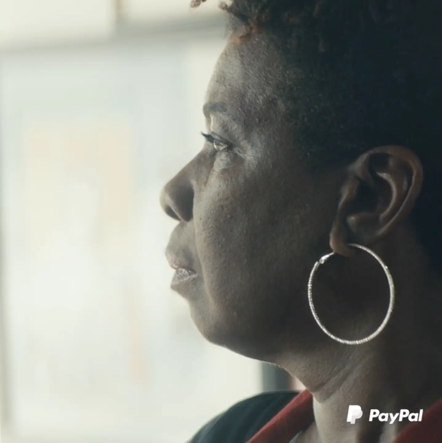 A woman with hoop earrings, managing her PayPal account, looks out of a window.