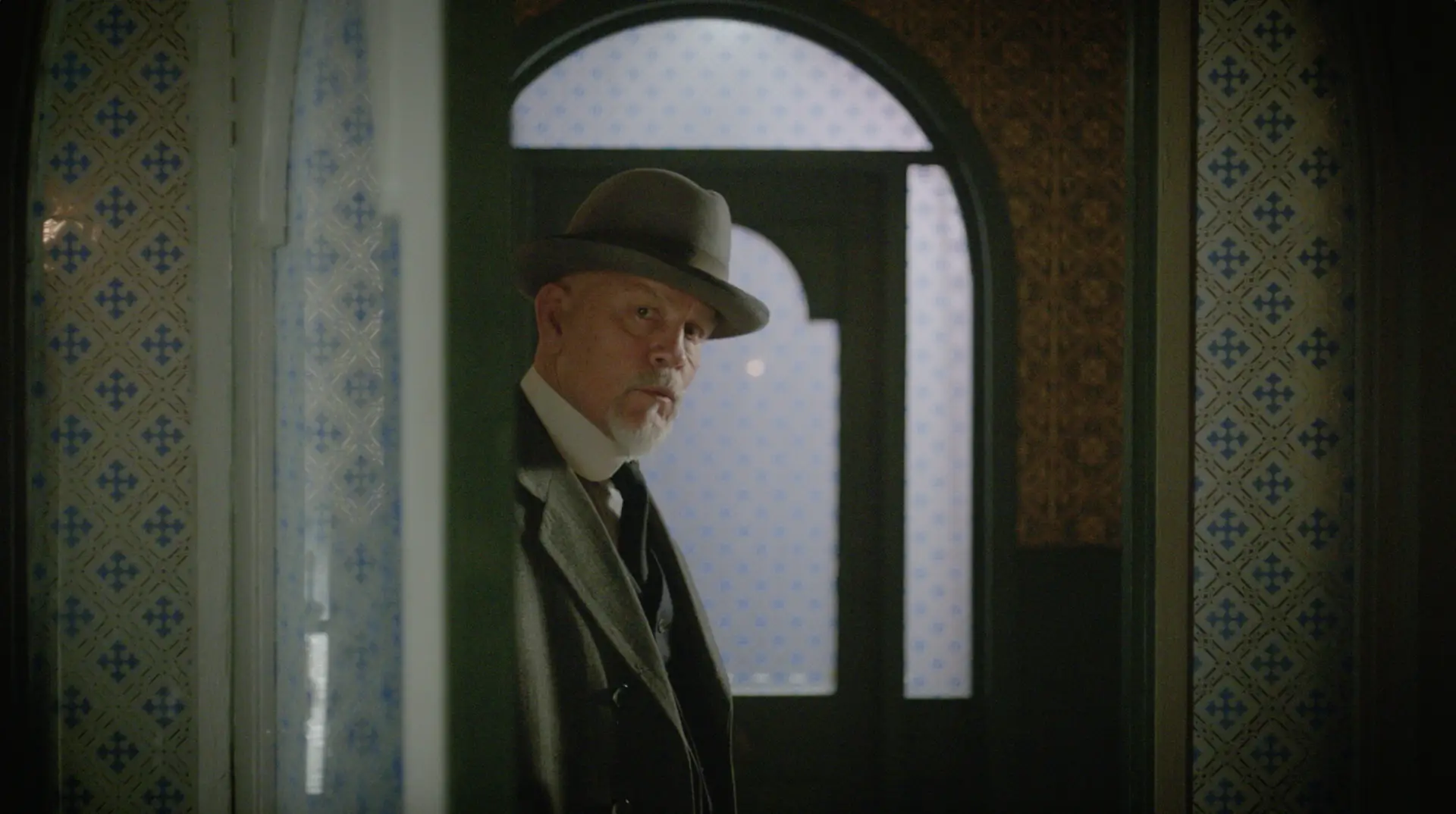 A man in a hat and coat is standing in a doorway, immersed in true crime work.