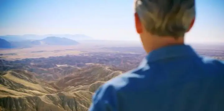 A man in a blue shirt is looking out over a desert, LIVE 2023.
