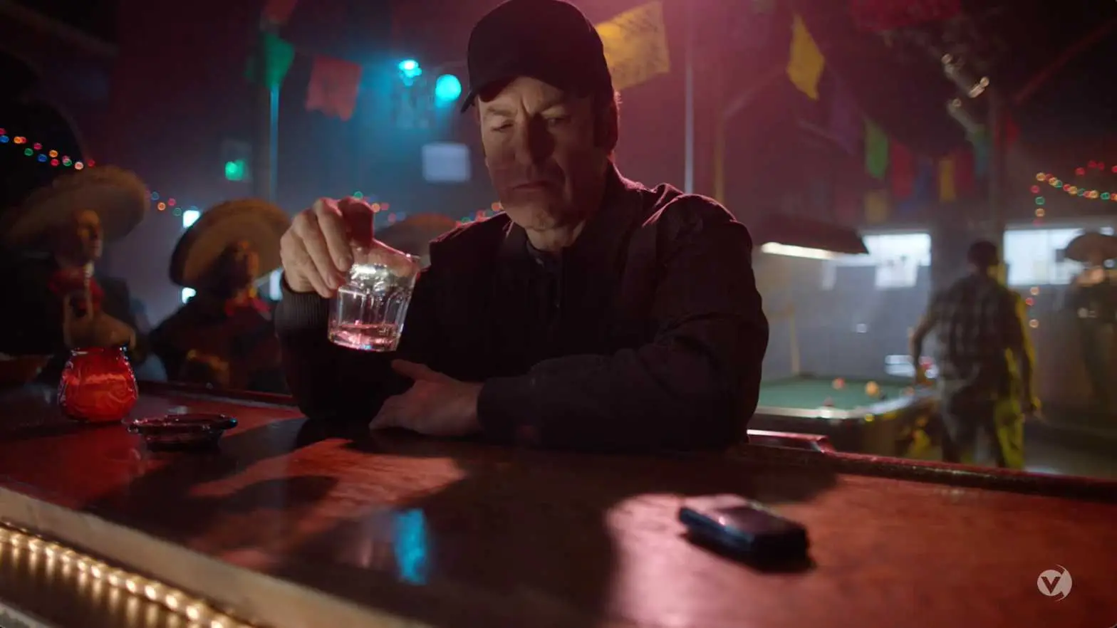 Man sitting at a bar with a somber expression holding a glass of alcohol, with festive decorations and people in the background, reminiscent of a scene from a content library.