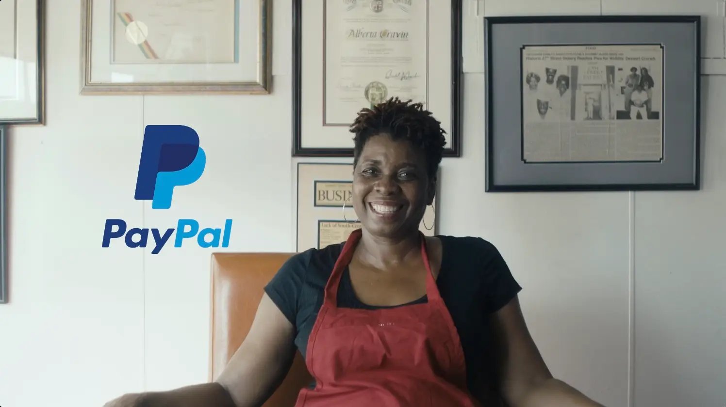 A smiling person wearing a red apron sitting in a room with framed certificates on the wall, displaying the PayPal logo on the left side, as part of a video content case study.
