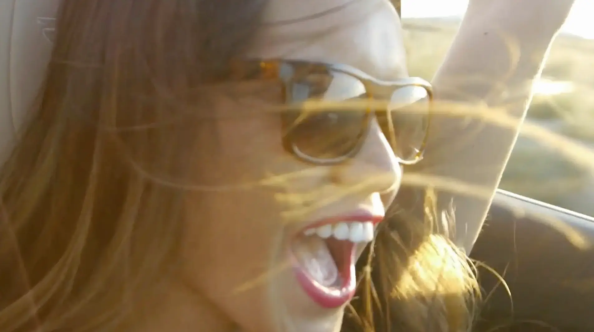 A joyful woman wearing sunglasses, shouting excitedly with her hair blowing in the wind, inside a moving car, heading to a presentation on Sales Tools.