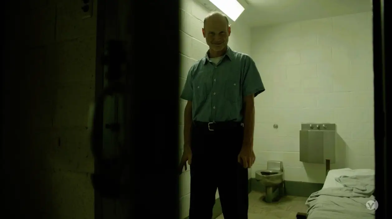 Man smiling in a dimly lit prison cell, standing by an open door with cinderblock walls and a bed to his side, resembling an unusual addition to a content library.